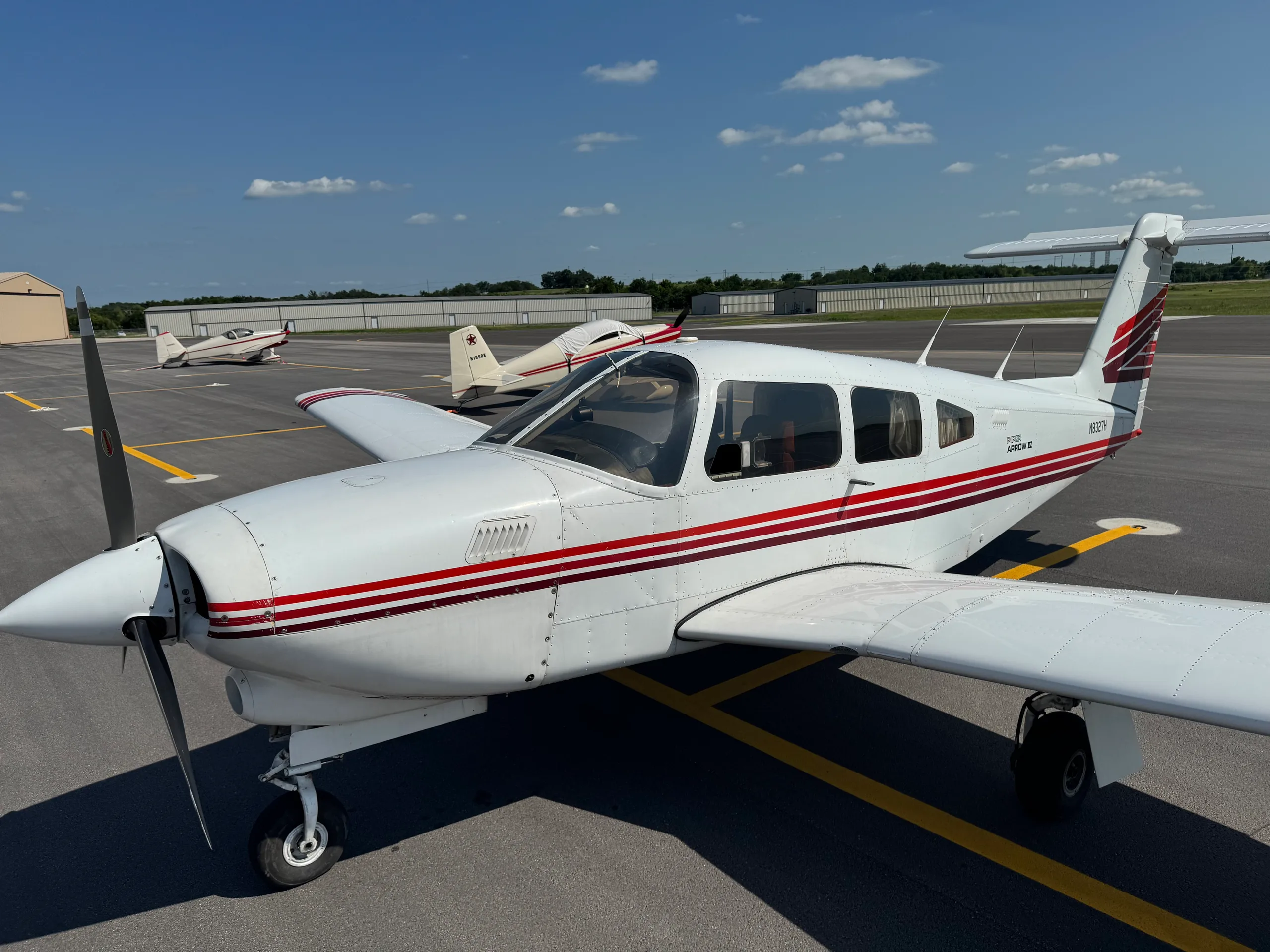 A white Piper Arrow II aircraft with red and brown stripes parked on an asphalt tarmac, viewed from the front left.