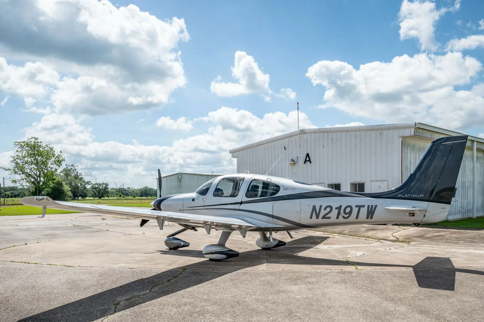 A side view of the Cirrus SR22T GTS Platinum aircraft N219TW parked on an airfield with a hangar and trees in the distance.