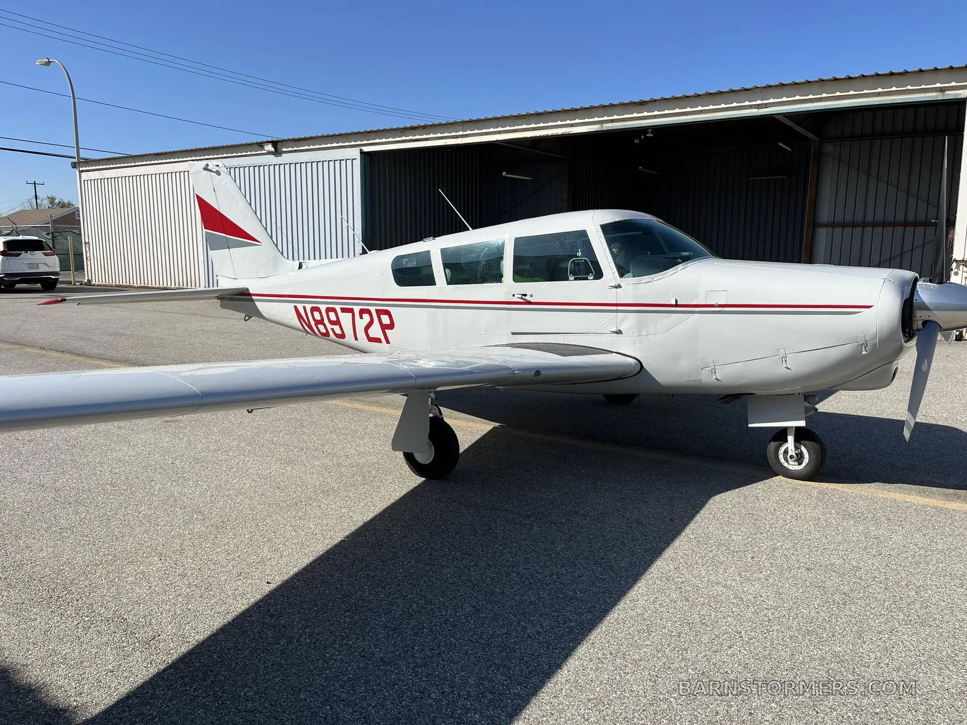 A white single-engine aircraft with red and grey stripes (N8972P) is parked on an asphalt ramp in front of an open hangar.
