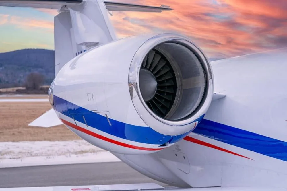 A close-up of the rear engine and tail of a white Learjet 45 with blue and red stripes, under a colorful sunset sky.