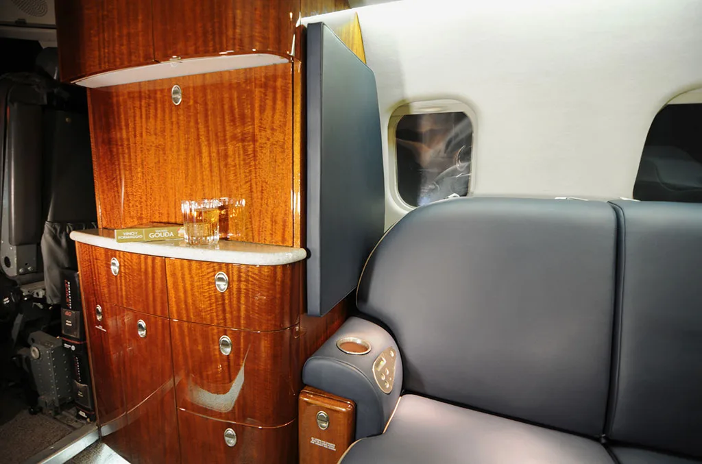 A close-up of the private jet interior showing a polished wood cabinet with drinks and a comfortable black leather sofa seat.