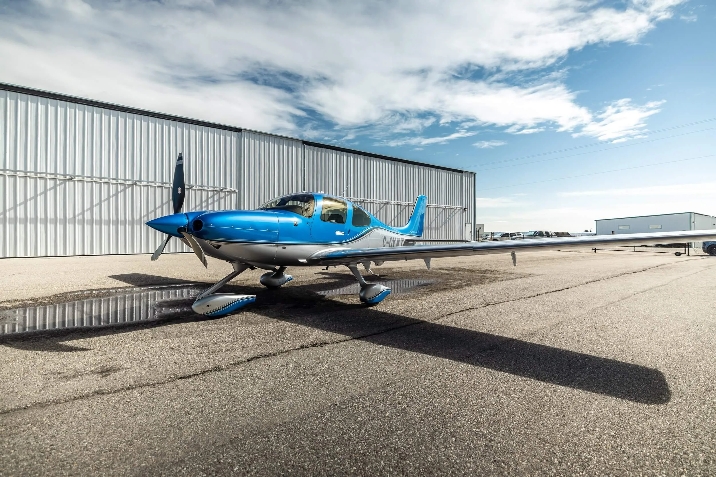 A blue and silver single-engine propeller aircraft, registration C-GYWT, parked on an asphalt tarmac next to a white hangar under a partly cloudy sky.