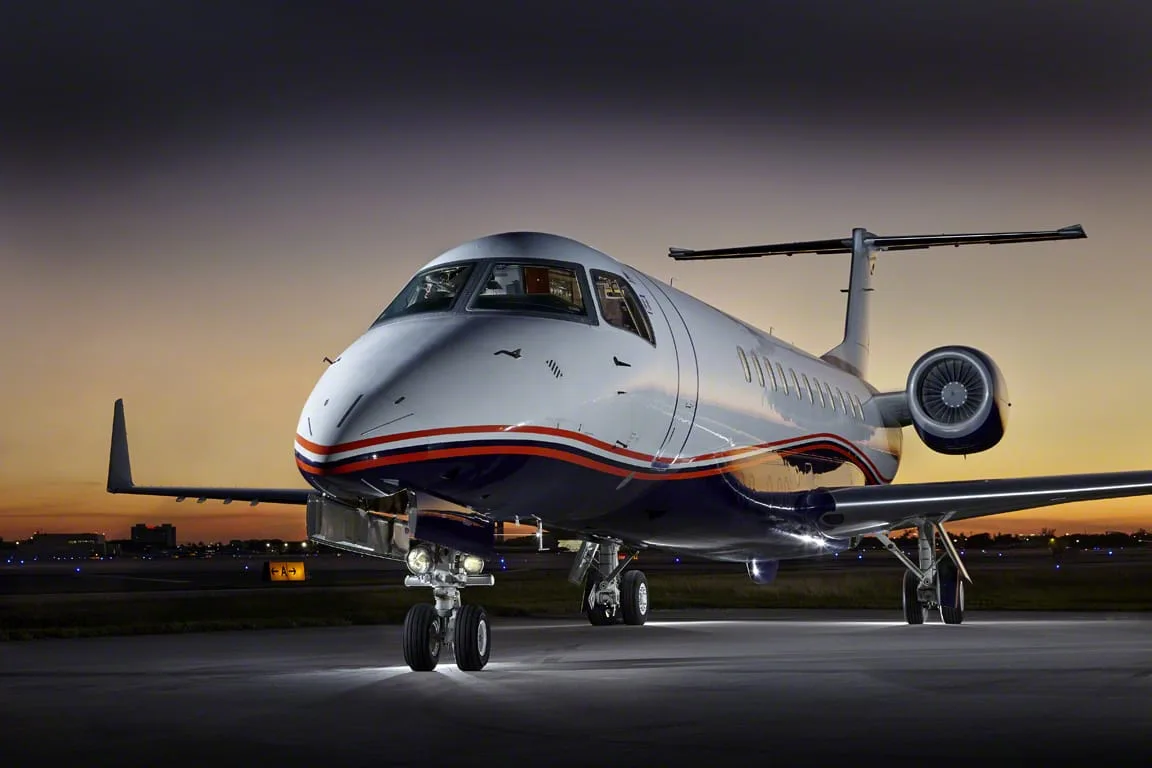 A white and blue Embraer Legacy 650 business jet parked on an airfield at dusk.