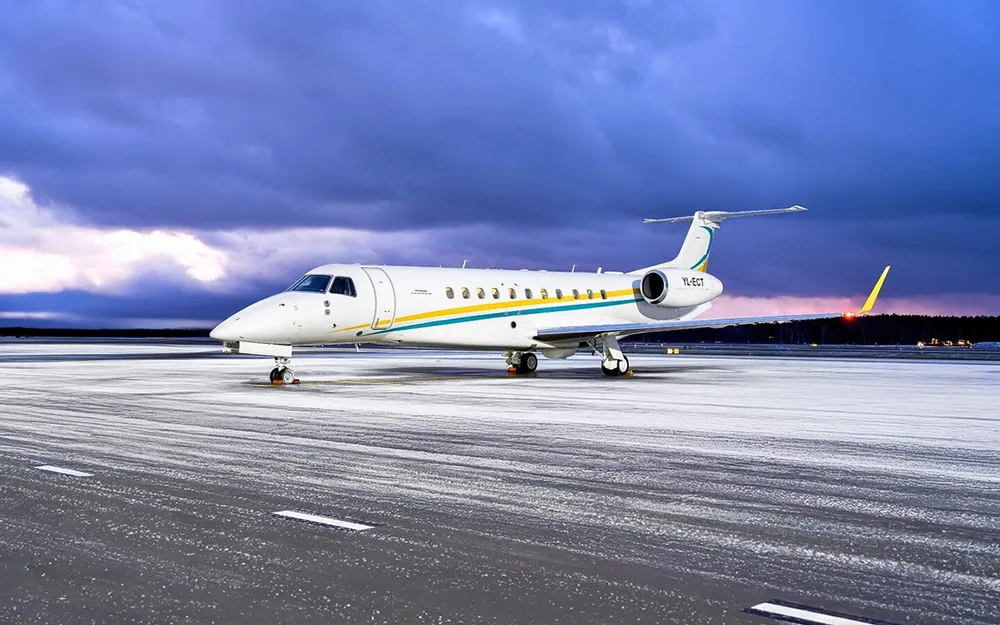 A white Embraer Legacy 600 business jet with blue and yellow stripes on a snowy tarmac under a dramatic sky.
