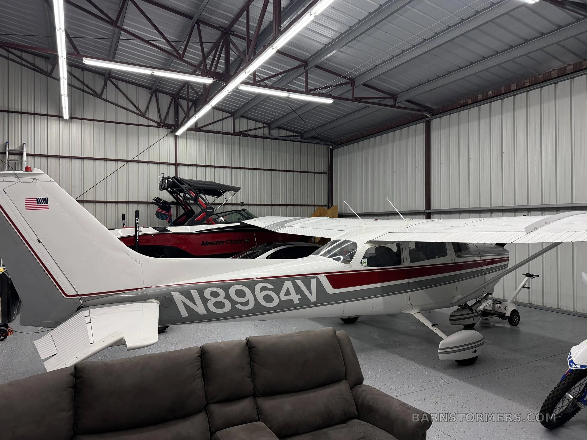 Rear view of a white and grey Cessna 172M with red stripes, registration N8964V, parked in a hangar with a brown couch in the foreground.