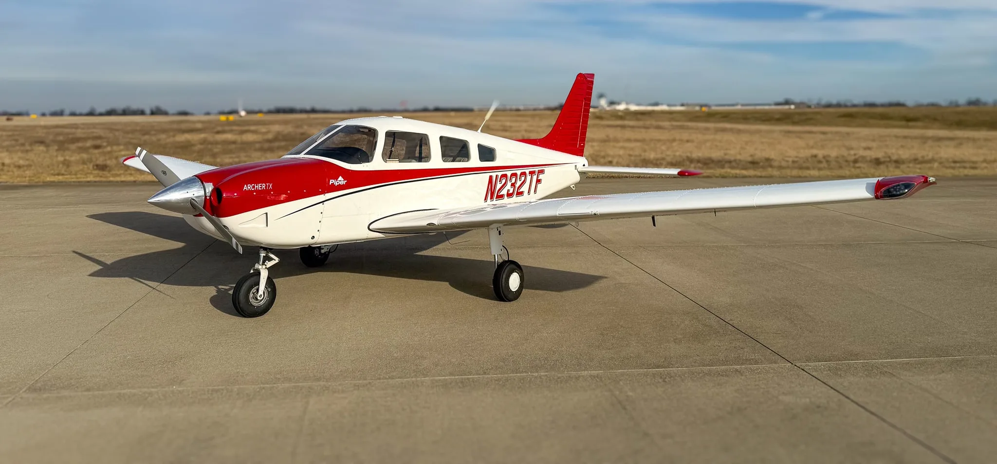 A white and red Piper Archer TX aircraft, tail number N232TF, parked on an airfield under a partly cloudy sky.