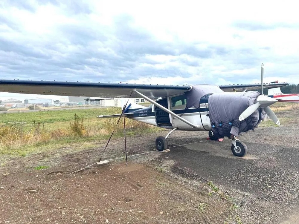 A white and blue Cessna 185 aircraft with engine and cockpit covers, parked on a gravel patch under a cloudy sky.