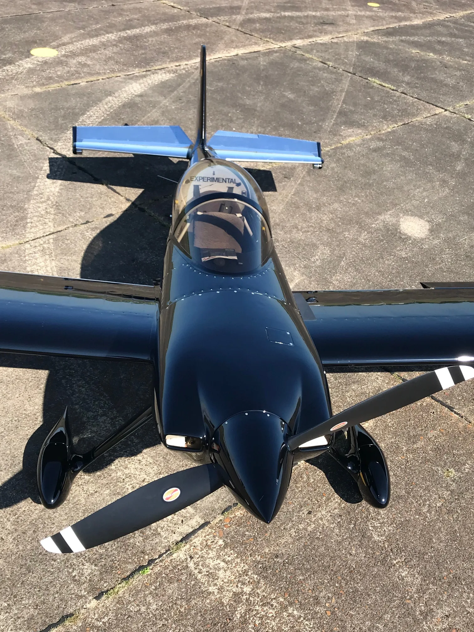 An overhead front view of the glossy black experimental aircraft with a two-bladed propeller on a concrete tarmac.