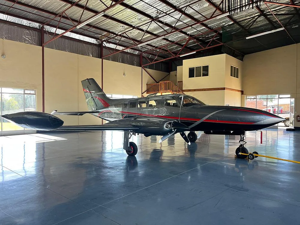 A dark grey twin-engine aircraft with red and silver accents, viewed from the front-right in a hangar.