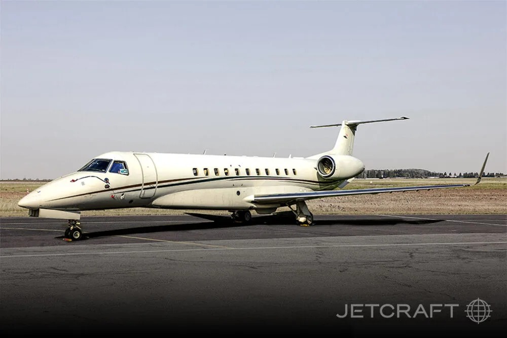A white Embraer Legacy business jet with a brown stripe parked on an asphalt runway under a clear sky.