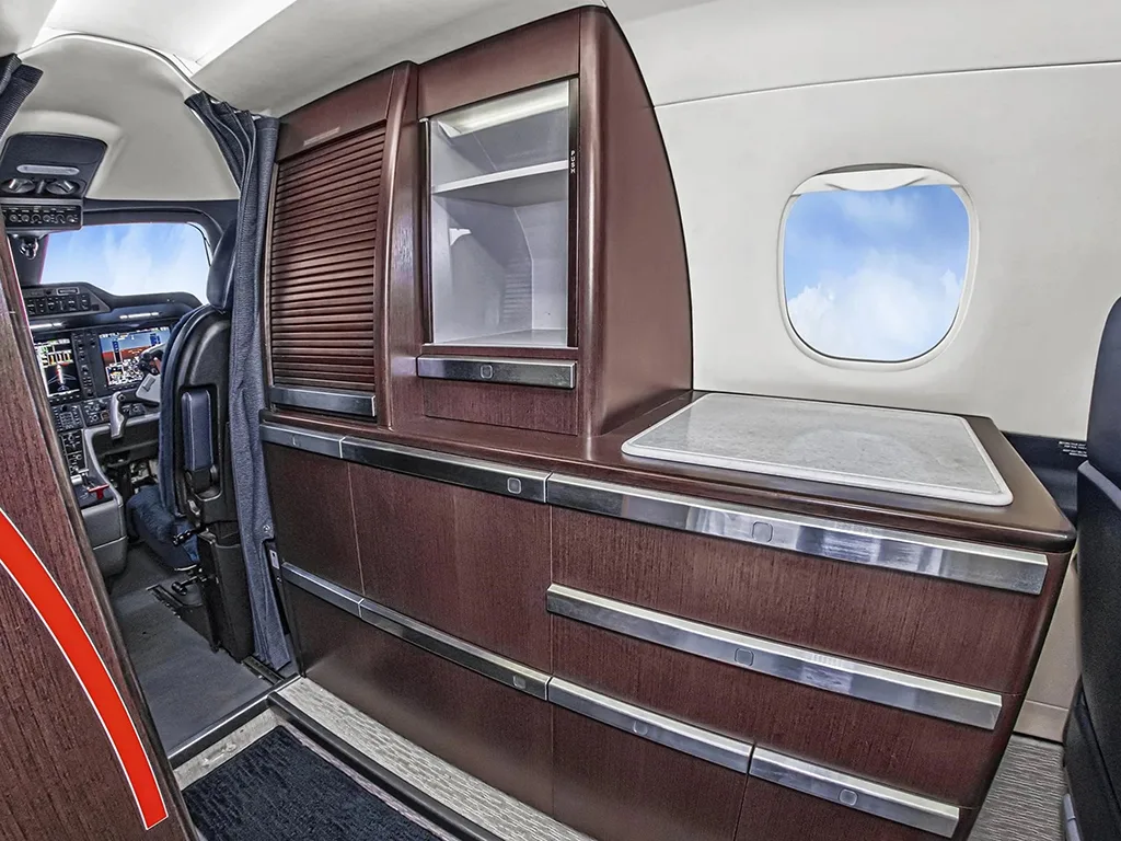 A dark wood galley cabinet with a countertop and storage is visible inside the aircraft cabin, with the cockpit in the background.