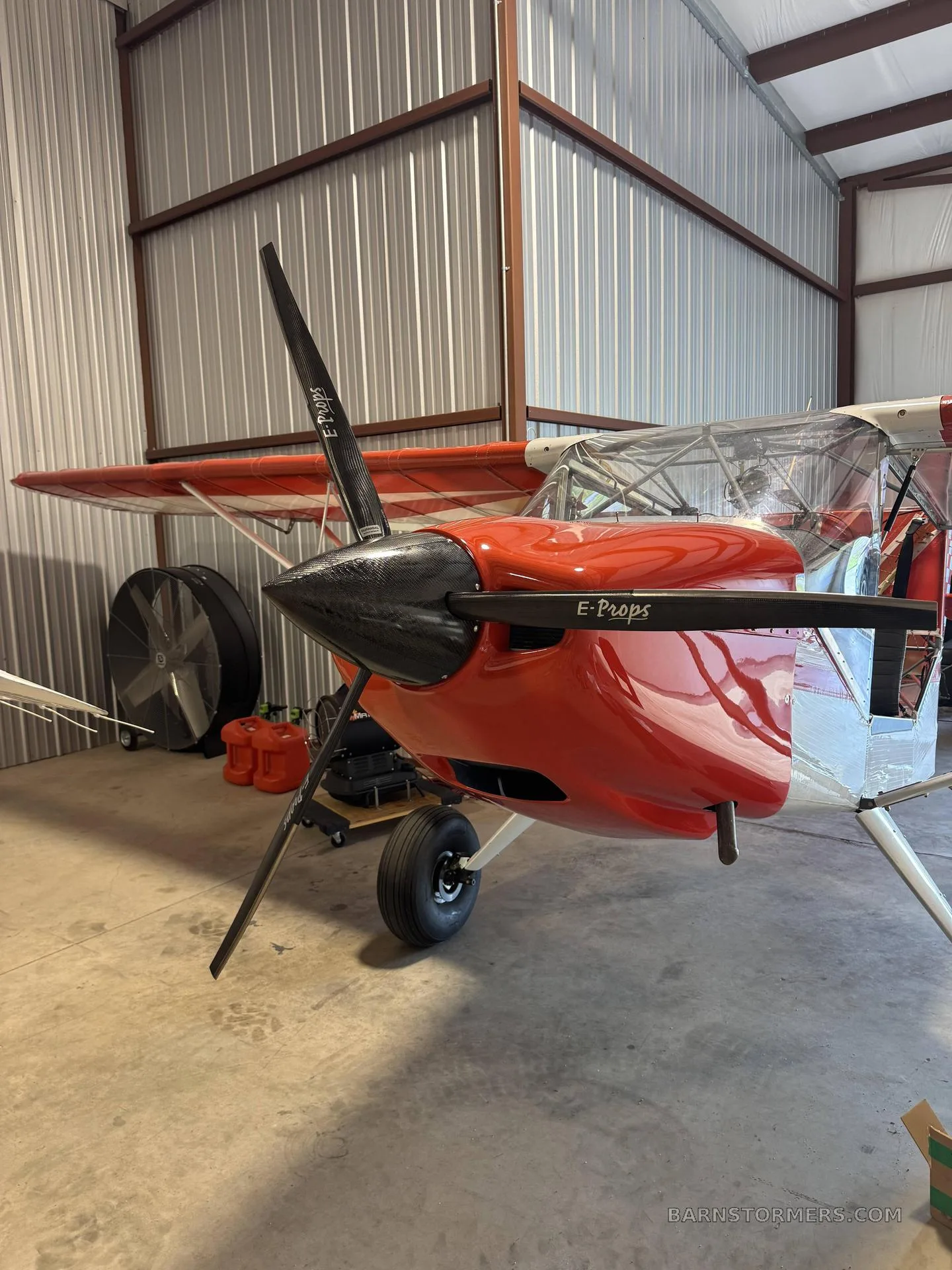 A red light sport aircraft with a black propeller, parked inside a metal hangar.
