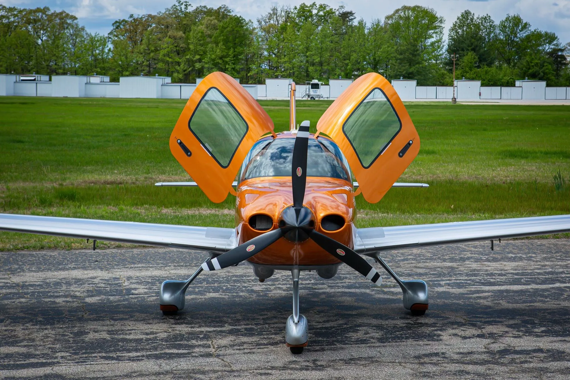 A front view of an orange and silver Cirrus SR20 aircraft with its gull-wing doors open, parked on an airfield.