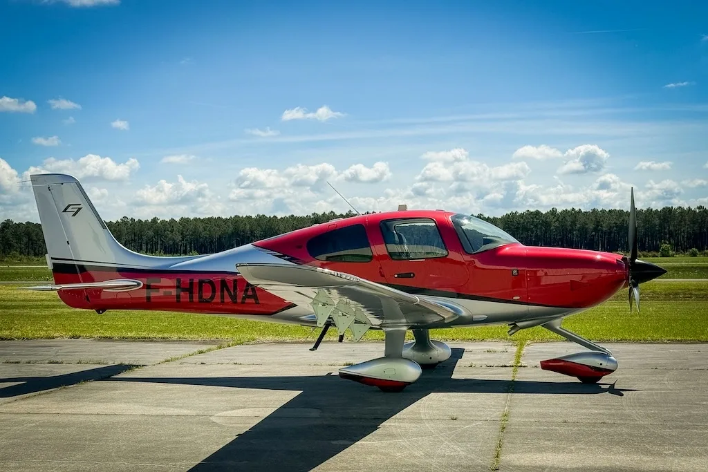 A red and silver Cirrus SR22 GTS (F-HDNA) aircraft parked on a tarmac with a forest in the background.