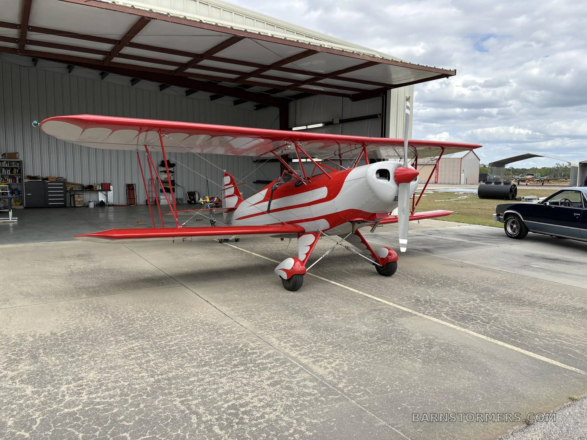 A red and white biplane with a radial engine is parked on concrete outside an open hangar.