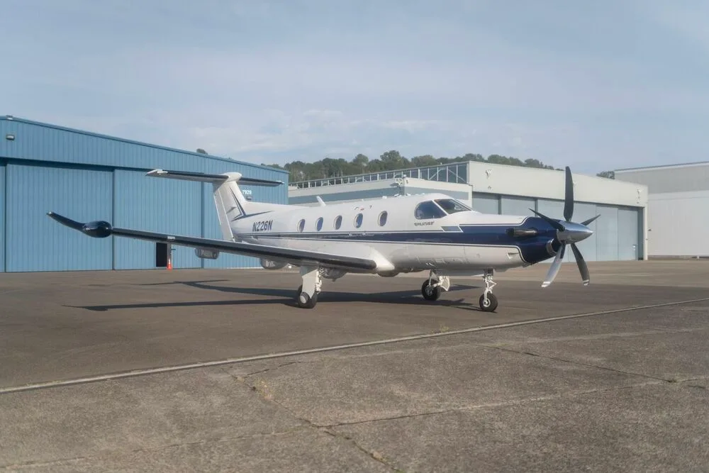 A white and blue Pilatus PC-12 aircraft with registration N226N parked on an tarmac with hangars in the background.