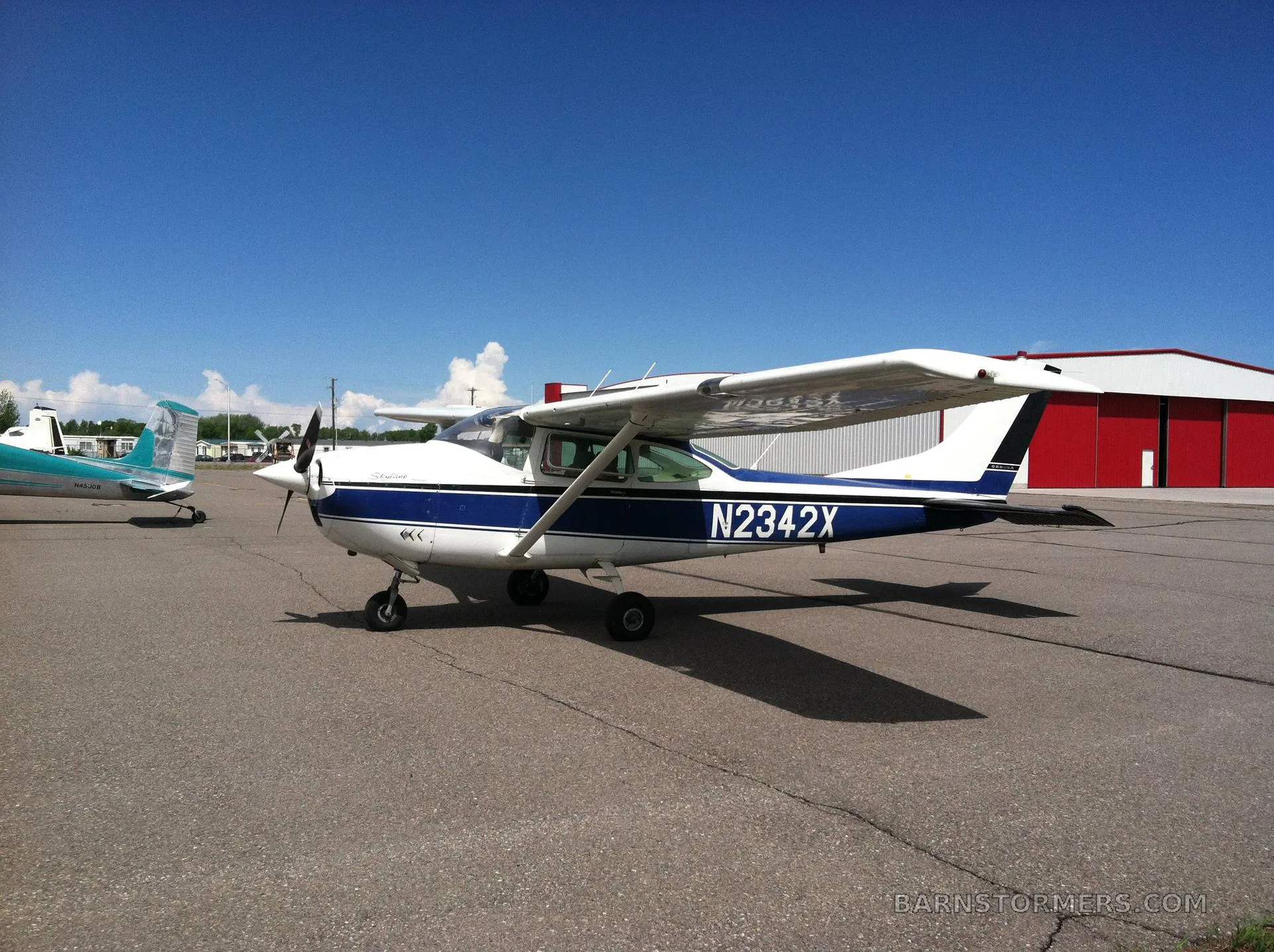 A white and blue Cessna 182 Skylane (N2342X) parked on an asphalt ramp next to a red hangar under a blue sky.