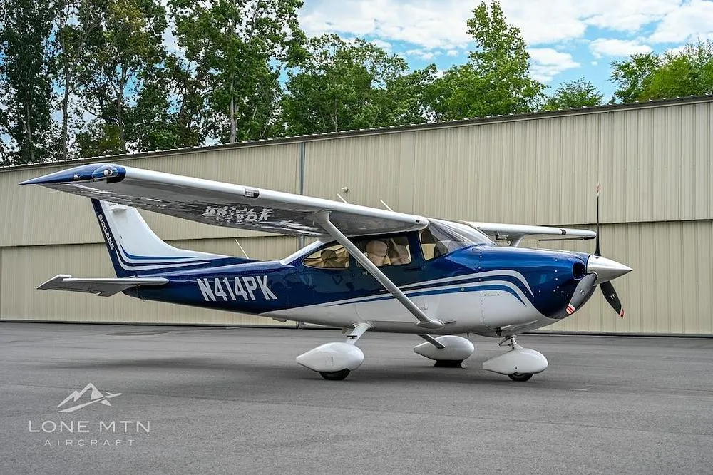A blue and white Cessna Skylane (N414PK) is parked on an asphalt apron in front of a hangar.