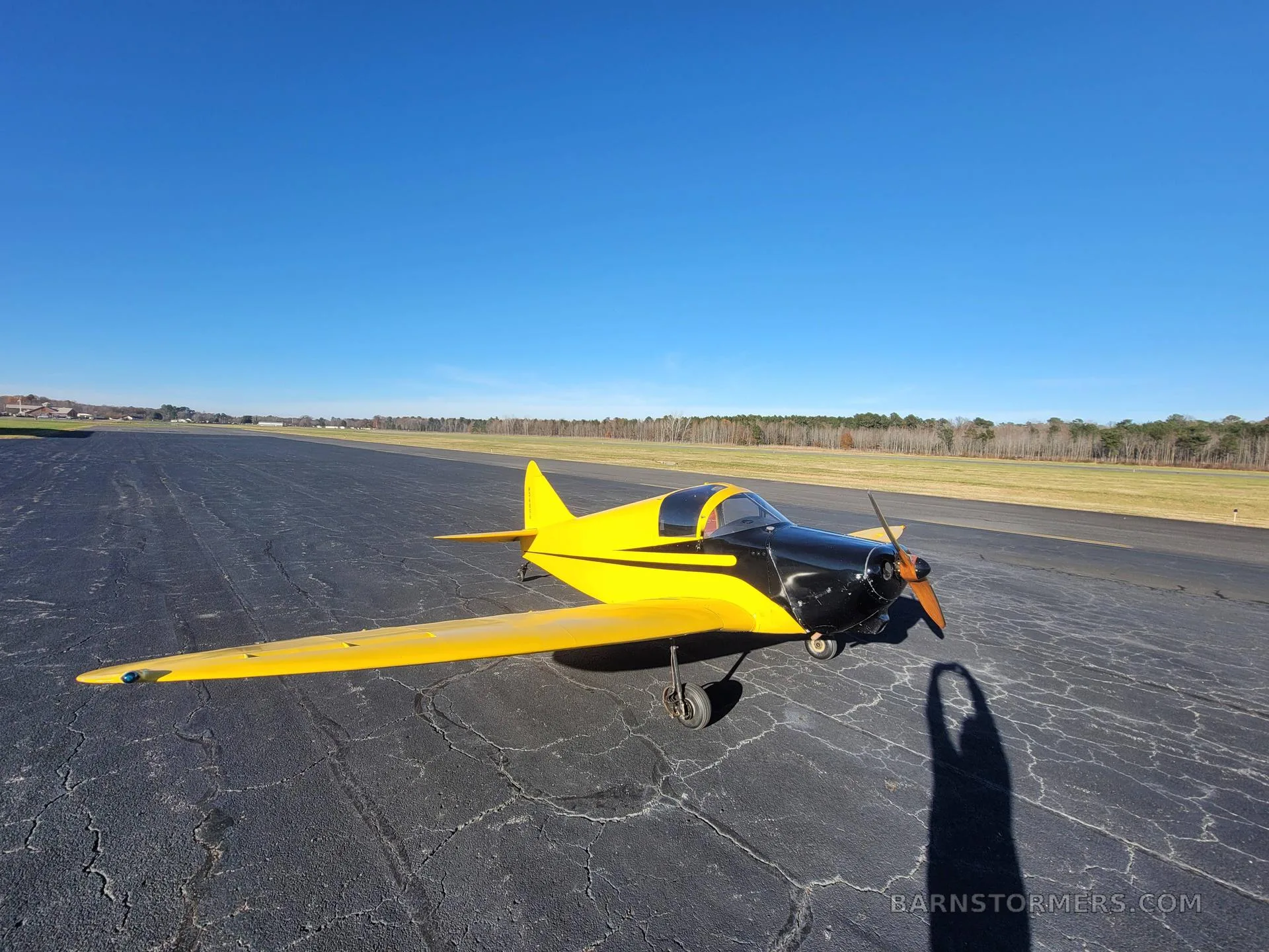 A yellow and black single-engine aircraft with a propeller, viewed from the front-right, on an asphalt surface under a clear blue sky.
