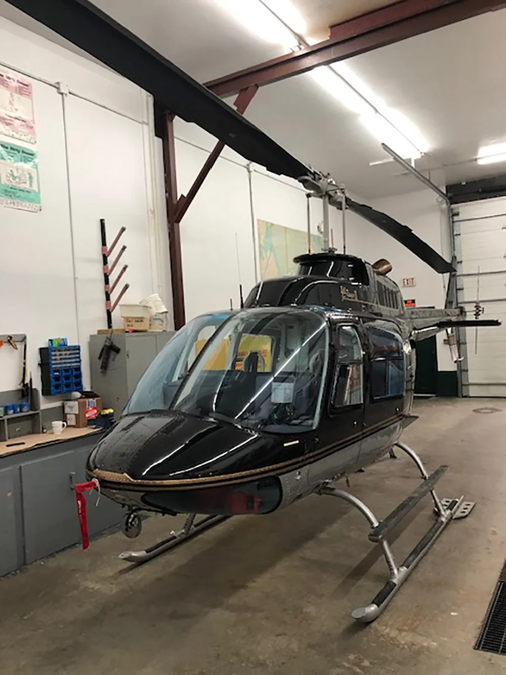 A black helicopter with silver landing skids is parked inside a well-lit hangar.