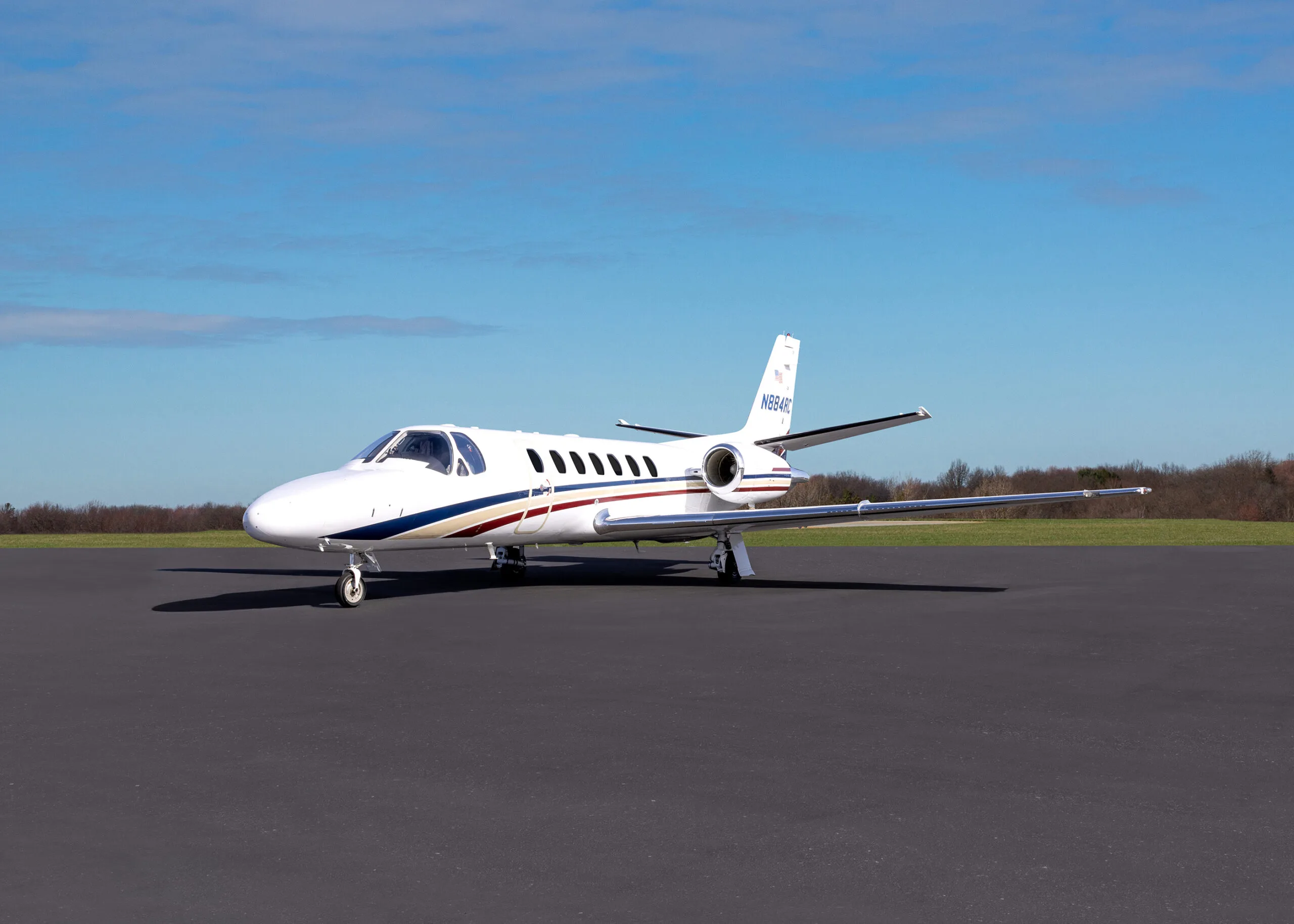 A white Cessna Citation business jet with blue, red, and gold stripes is parked on an asphalt tarmac under a clear blue sky.