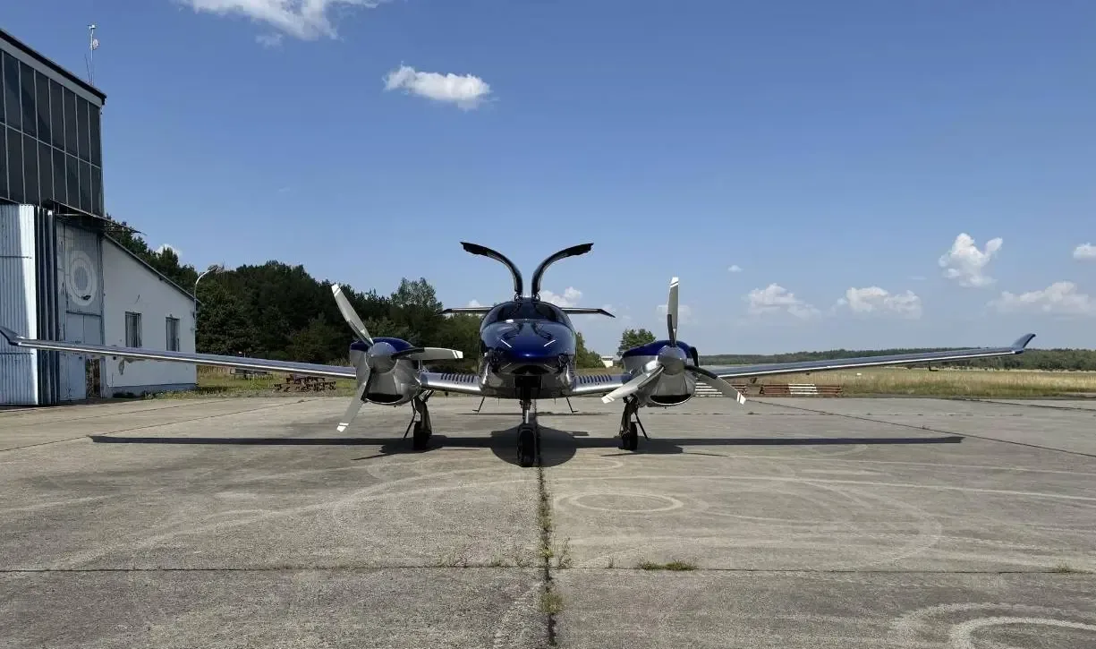 A front view of a blue and silver Diamond DA62 aircraft with its gull-wing doors open on a sunny tarmac.