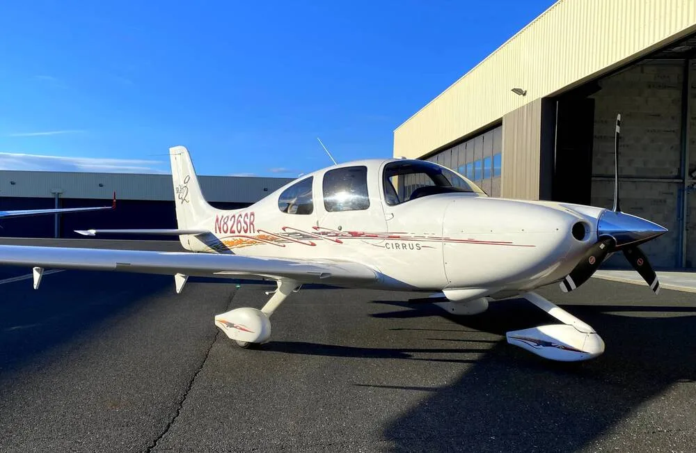 A white Cirrus SR22 aircraft with red and gold accent stripes, registration N826SR, parked on an airfield next to a hangar.