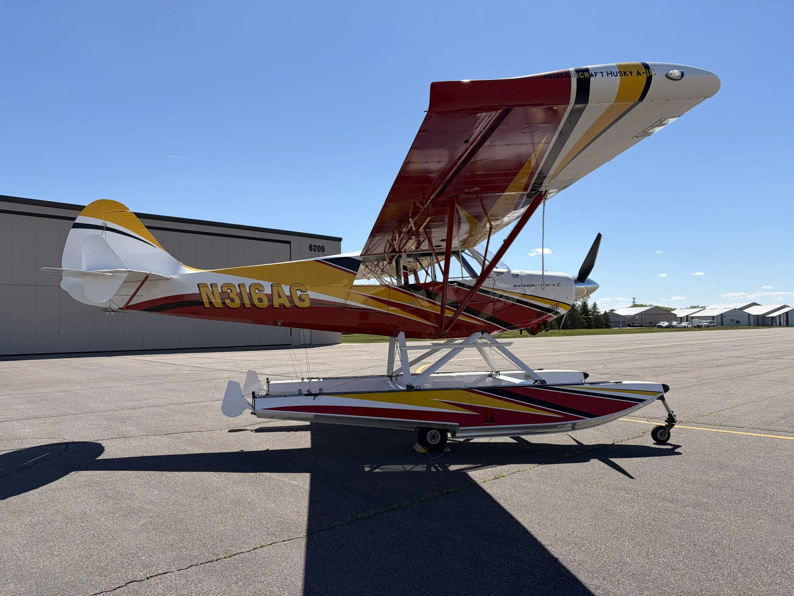 A side view of the red, yellow, and white Aviat Aircraft Husky A-1C seaplane, N316AG, parked on an airfield.