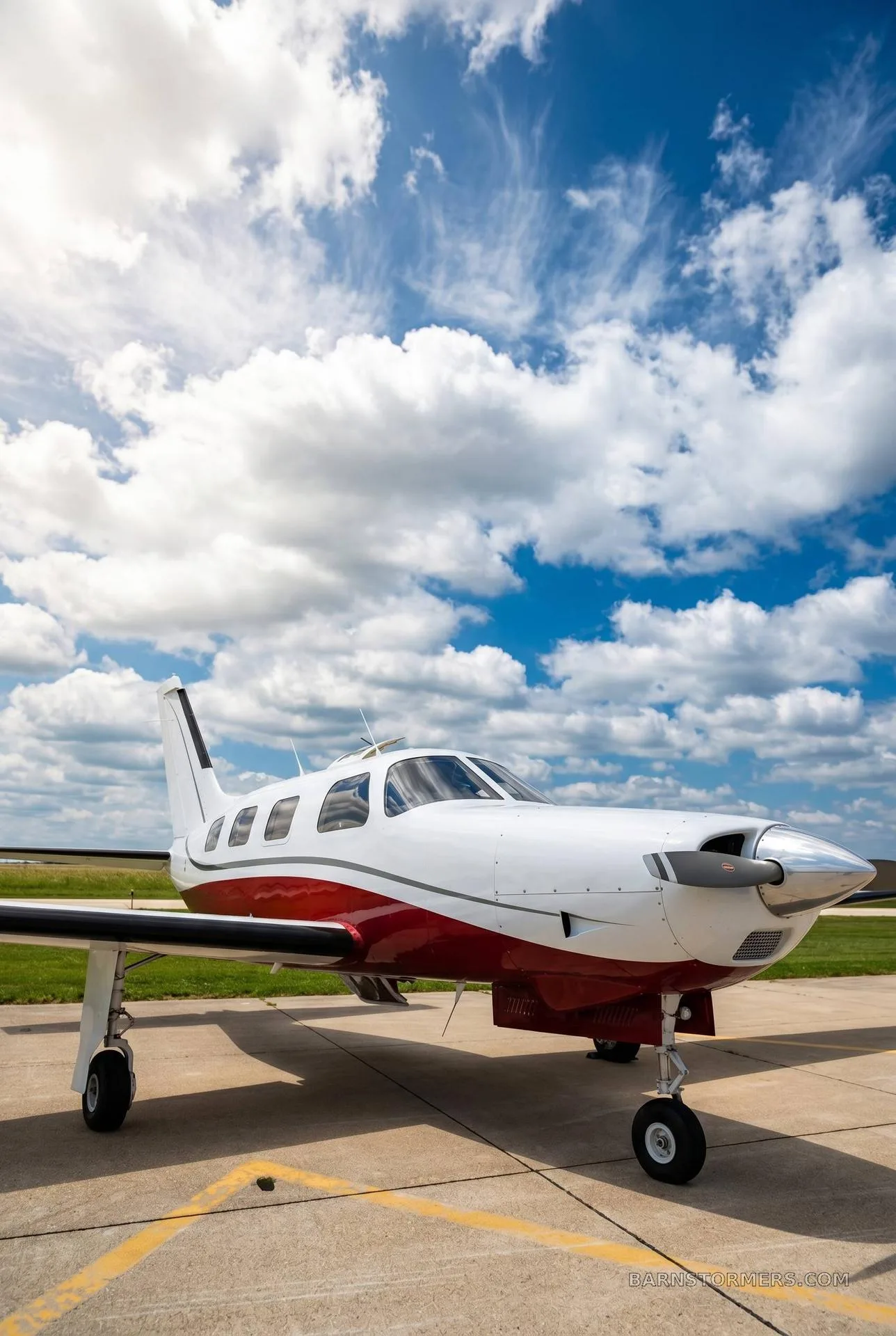 A white and red single-engine aircraft with a propeller is parked on a tarmac under a bright blue sky with fluffy white clouds.