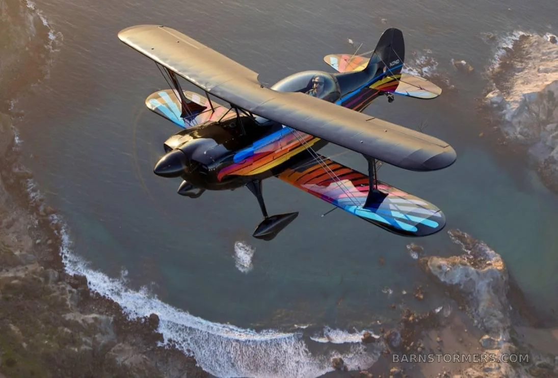 A black biplane with a colorful red, orange, and blue paint scheme on its wings and tail, flying over a rocky coastline with ocean waves.