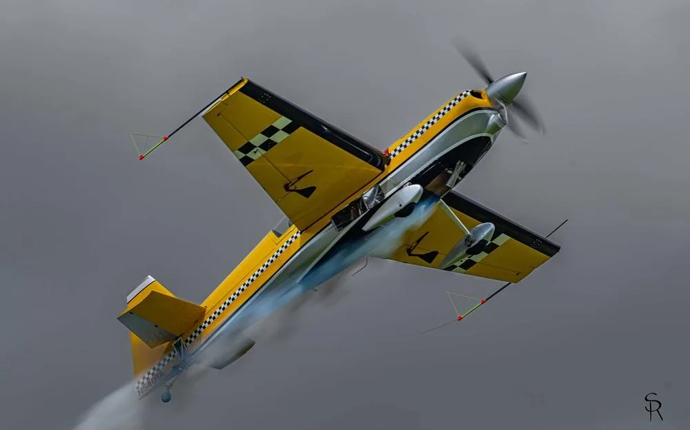 A yellow aerobatic aircraft with checkerboard wings and fuselage performs a maneuver, leaving a smoke trail.