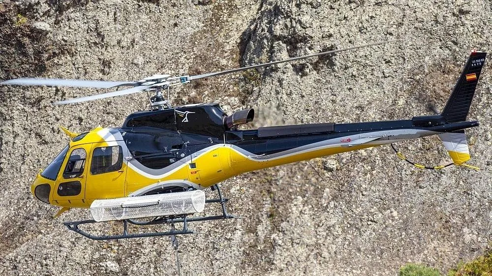 A yellow and black Airbus H125 helicopter with external equipment flying against a rocky background.