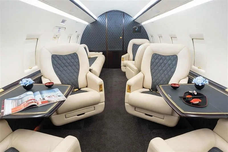The luxurious interior cabin of a business jet with cream and dark grey quilted seats, showing tables set with sushi and magazines.