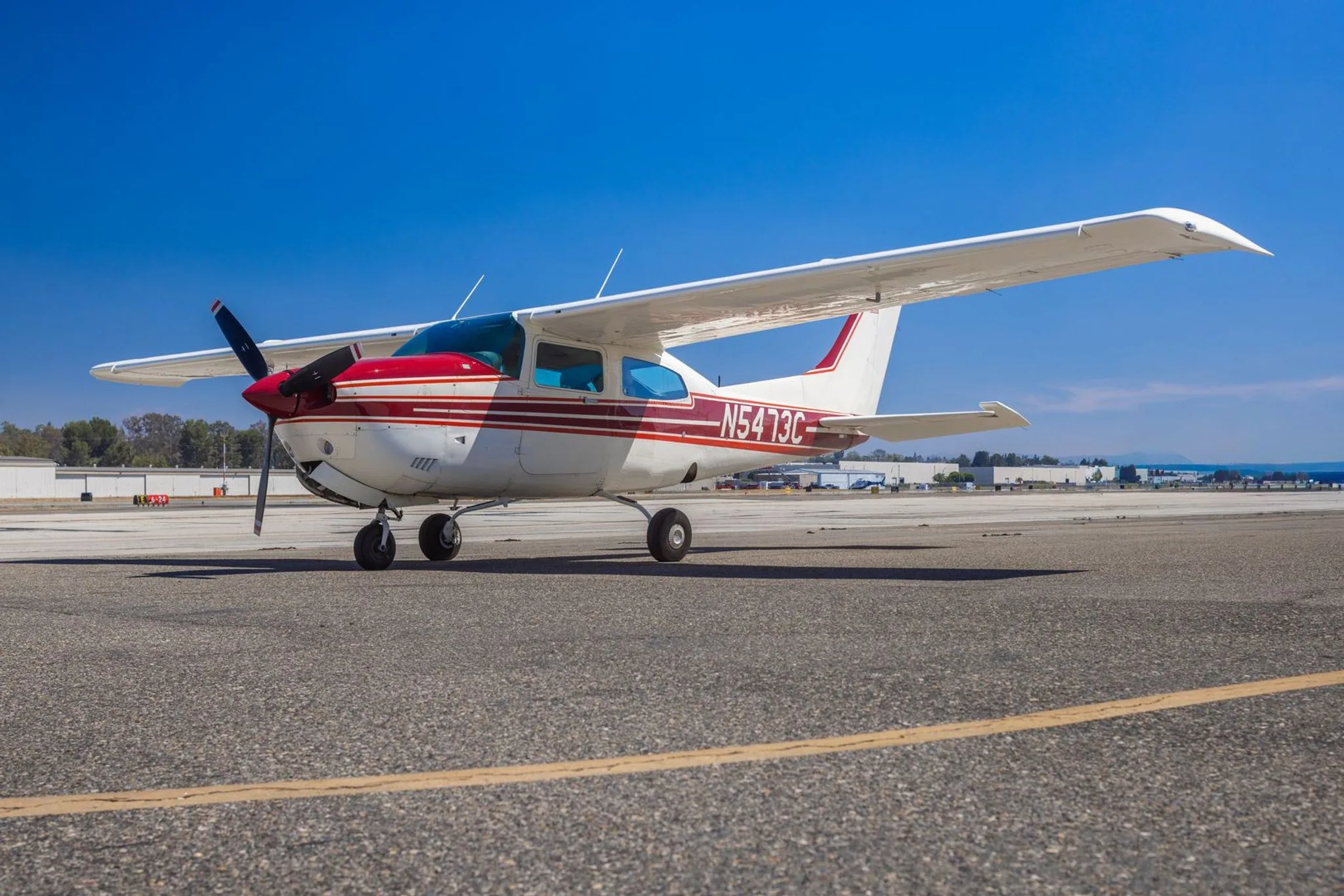 A white and red Cessna 210 Centurion aircraft, tail number N5473C, parked on an asphalt tarmac under a clear blue sky.