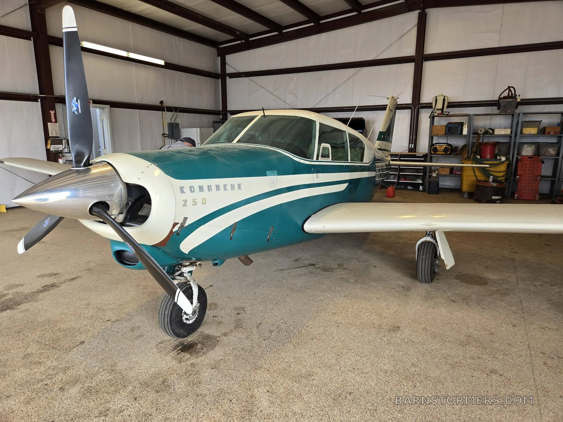 A front-left view of a white and teal Piper Comanche 250 aircraft with its propeller, parked inside a hangar.
