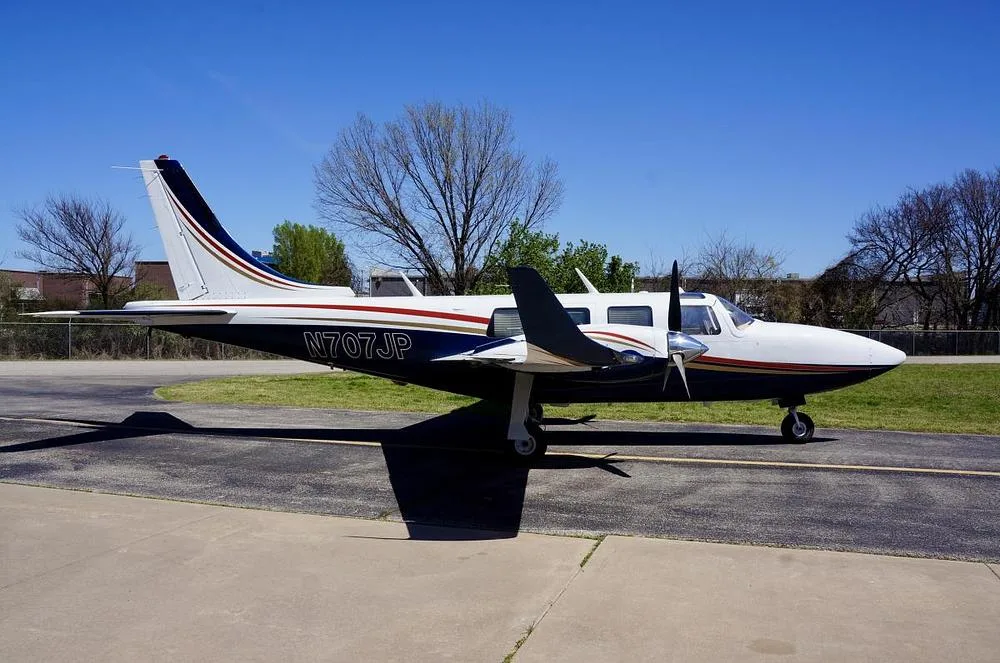 A side view of a white and dark blue twin-engine propeller aircraft with gold and red stripes, parked on an asphalt ramp under a clear blue sky.