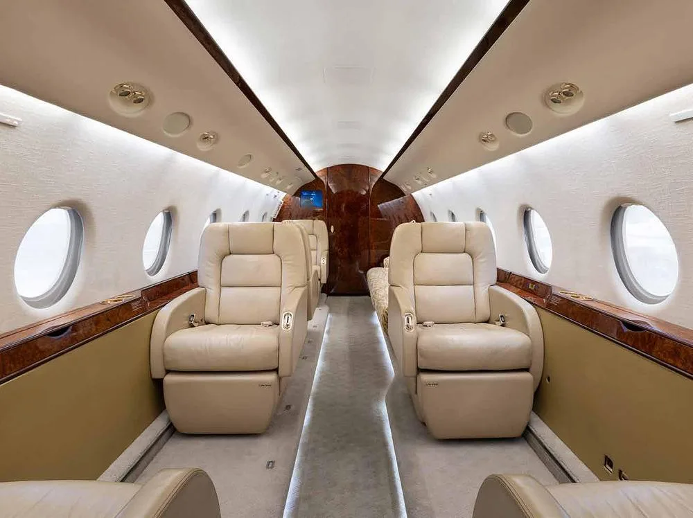 The luxurious interior cabin of a private jet with beige leather seats, wood paneling, and large oval windows.