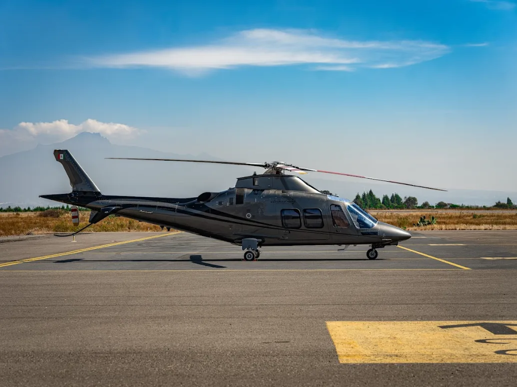 A grey AgustaWestland AW109 GrandNew helicopter is parked on an airfield with distant mountains under a partly cloudy sky.