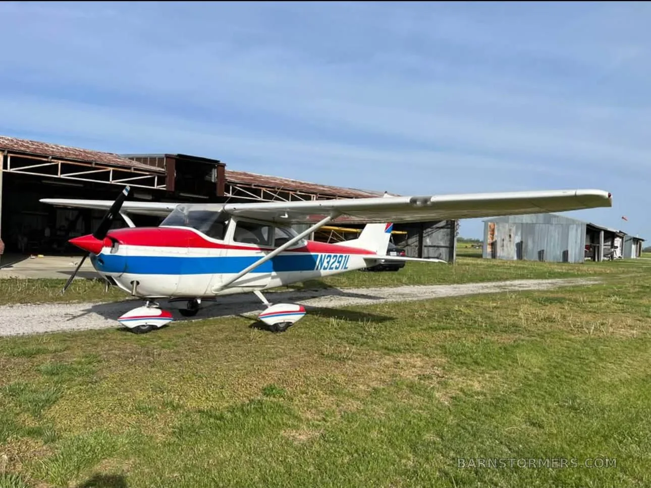 A white, red, and blue Cessna-style aircraft with registration N3291L parked on a gravel path next to hangars.