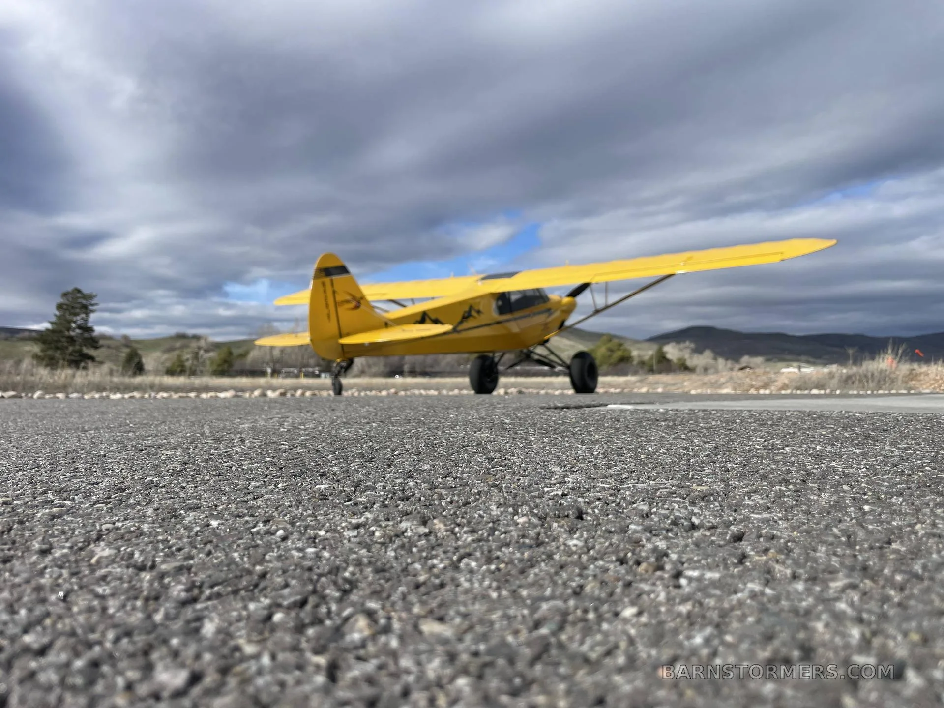 A very low-angle shot of a yellow CubCrafters Carbon Cub EX-2 with mountain decals, parked on an asphalt runway under a cloudy sky.