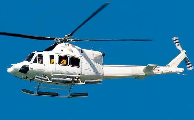 A white helicopter with an Italian flag on its tail flies against a clear blue sky.