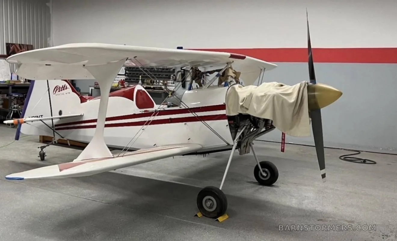 A white and red Pitts Special biplane with its engine exposed and propeller covered, parked in a hangar.