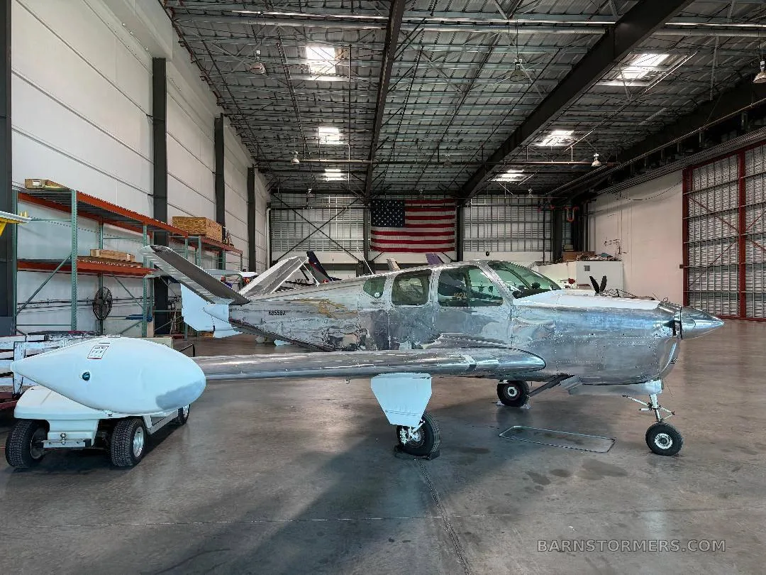 A bare metal, V-tail aircraft (N8558A) is parked in a hangar with an American flag in the background.