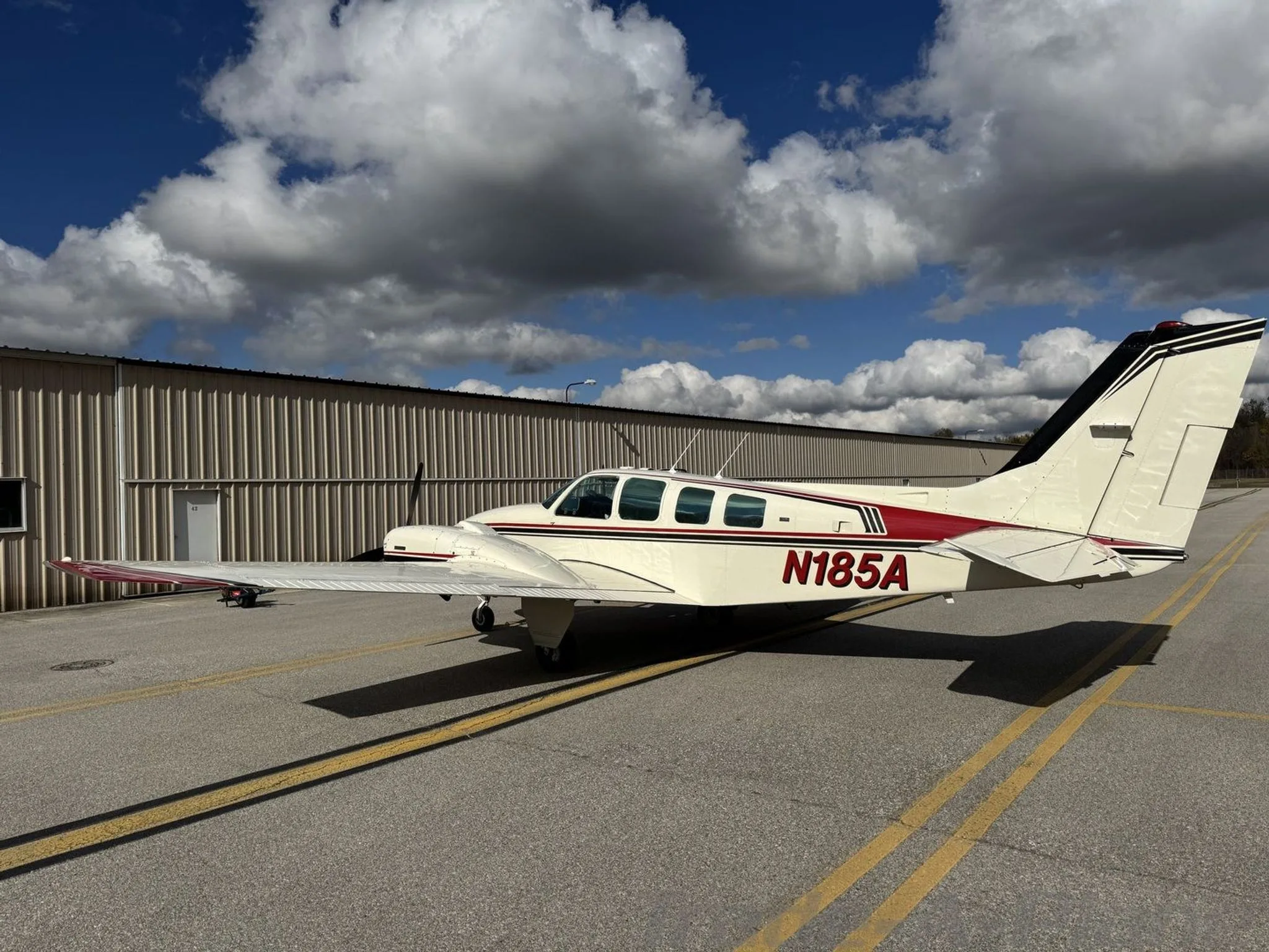 A side-rear view of the N185A twin-engine aircraft, white with red and black stripes, parked on an airport tarmac under a cloudy sky.