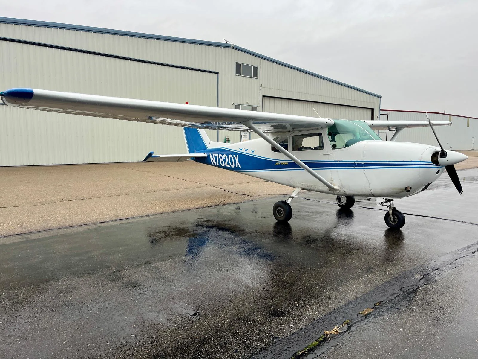 A white Cessna 172 Skyhawk with blue stripes and tail, registration N7820X, parked on a wet tarmac in front of a hangar.