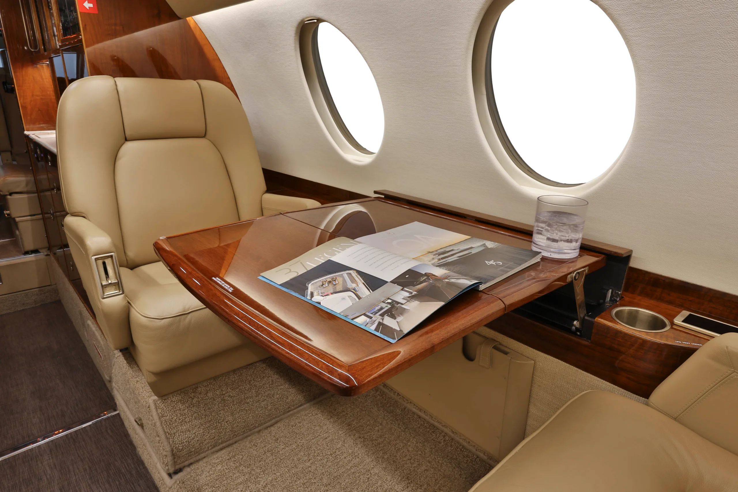 A close-up of a beige leather seat and a polished wooden table with a magazine and a glass of water inside a business jet cabin.
