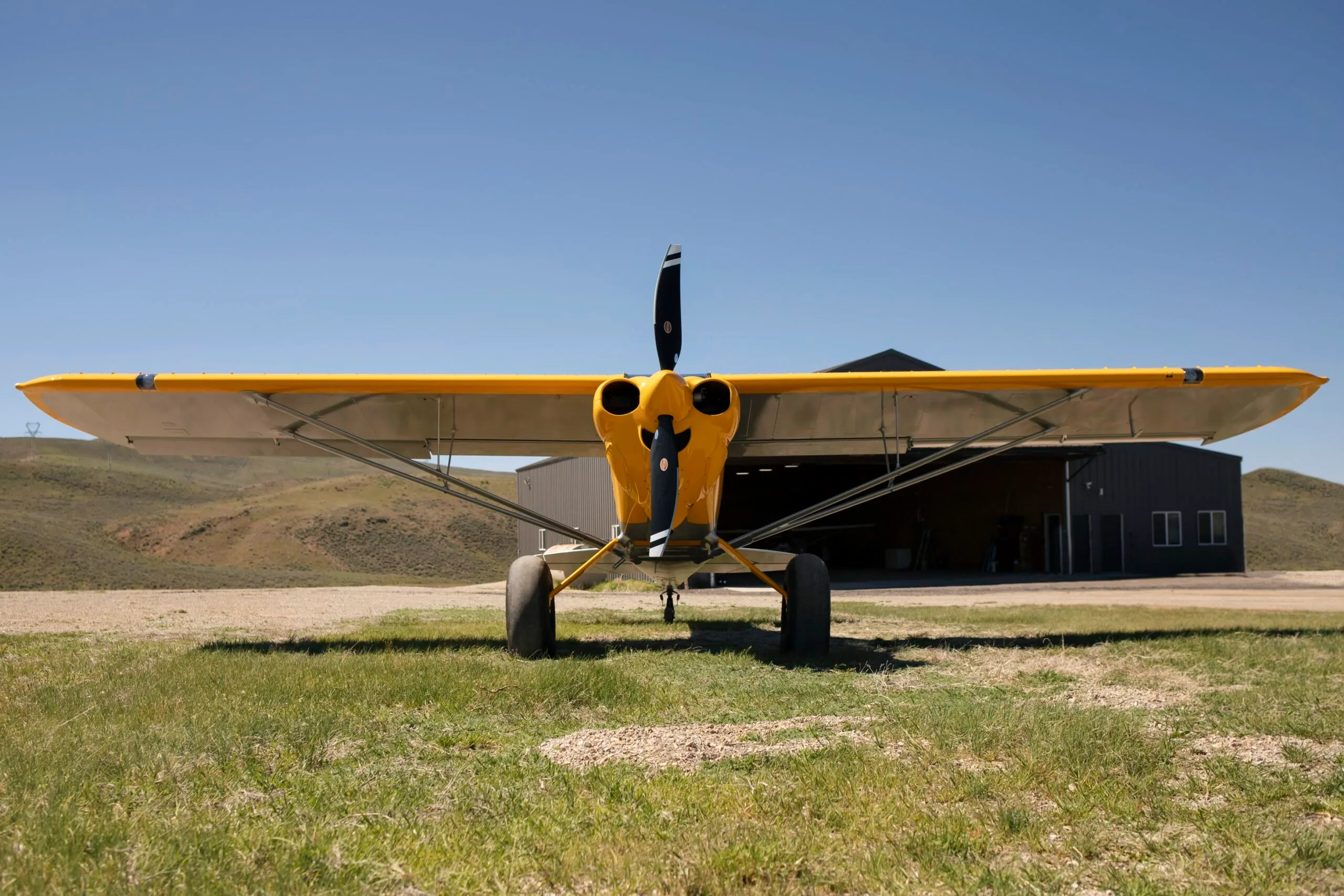 A front view of a yellow and silver CubCrafters XCub aircraft with a hangar in the background.