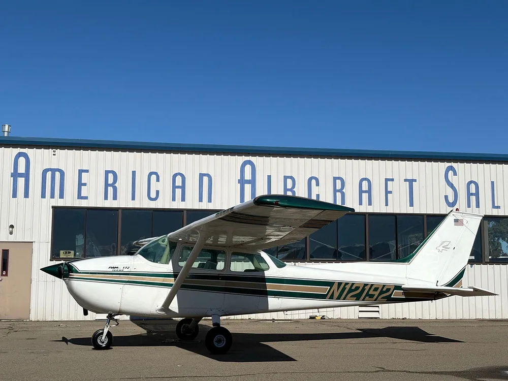 A white Cessna 172 with green and gold stripes parked on an airfield in front of a hangar, viewed from the left side.