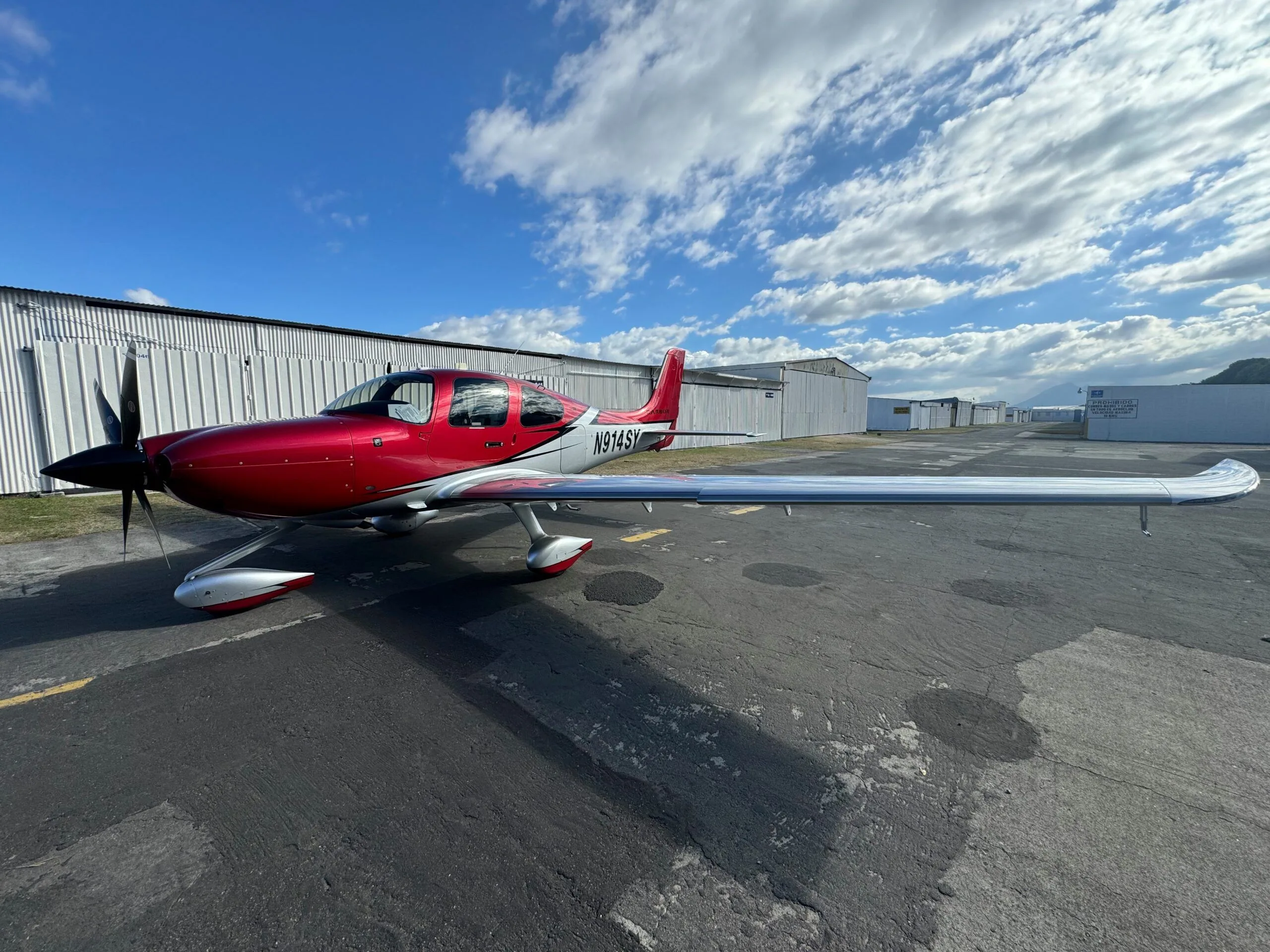 A red and silver Cirrus SR22 aircraft with registration N914SY parked on an asphalt apron under a partly cloudy sky.