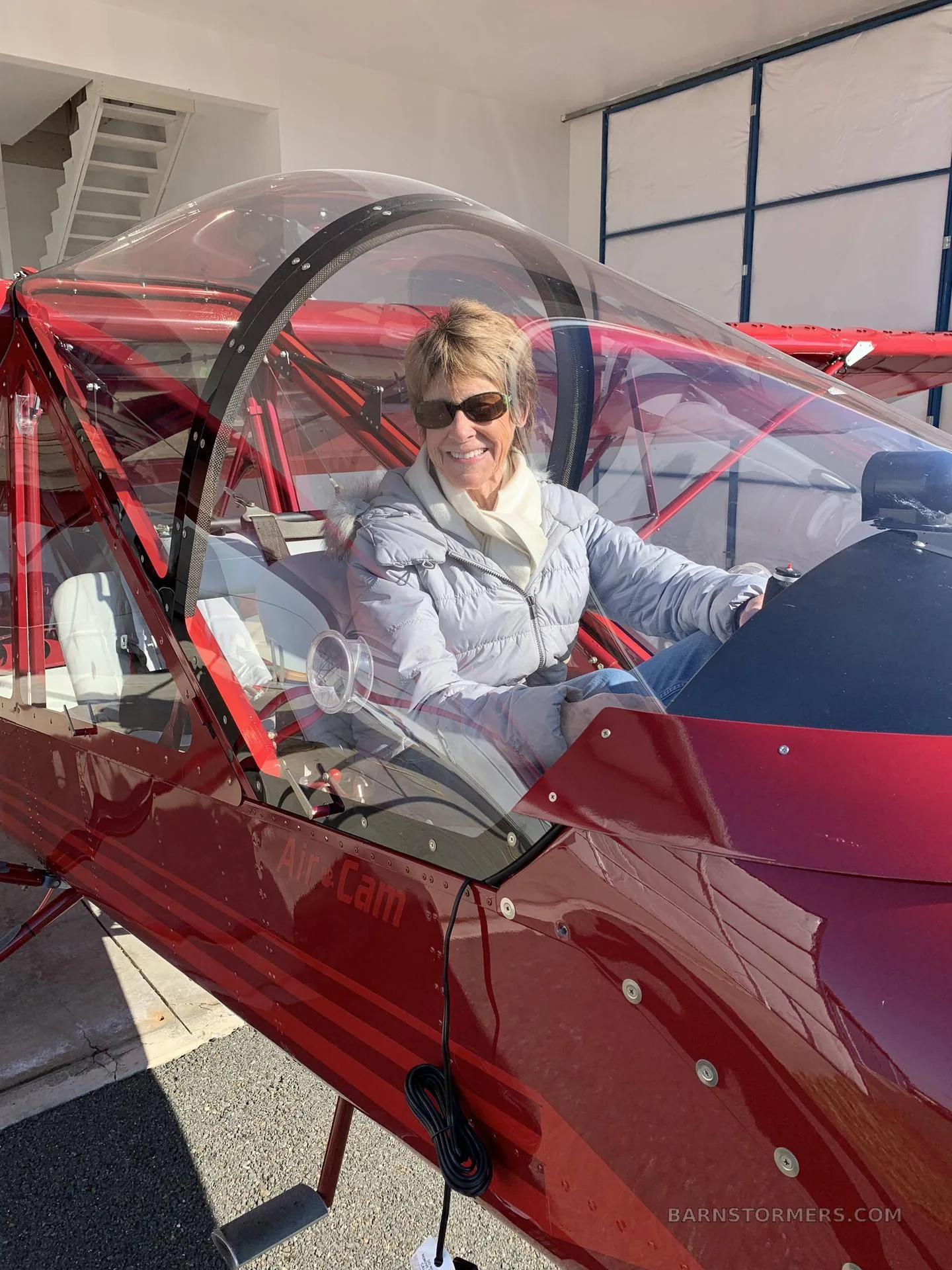 A woman in a grey jacket and sunglasses smiles from the cockpit of a red Air Cam aircraft.
